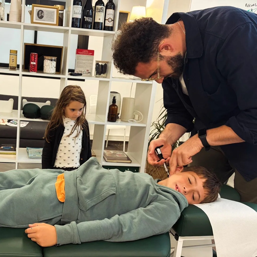 Chiropracteur qui ajuste un enfant allongé sur la table de consultation à la Clinique de la Colonne à Fenouillet, avec une petite fille qui observe, ambiance chaleureuse et rassurante