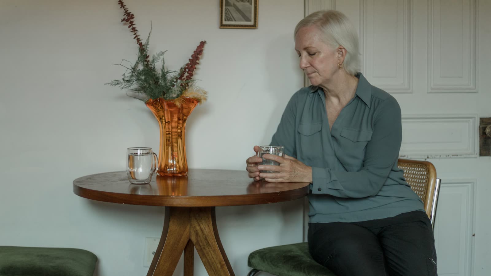 Femme d'une soixantaine d'années assise à une table, un verre d'eau entre les mains, l'air pensif