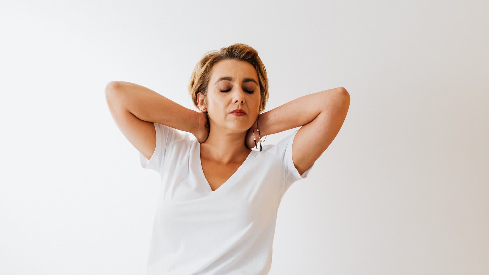 Femme assise à son bureau, les yeux fermés, qui relâche ses cervicales en les soutenant des deux mains