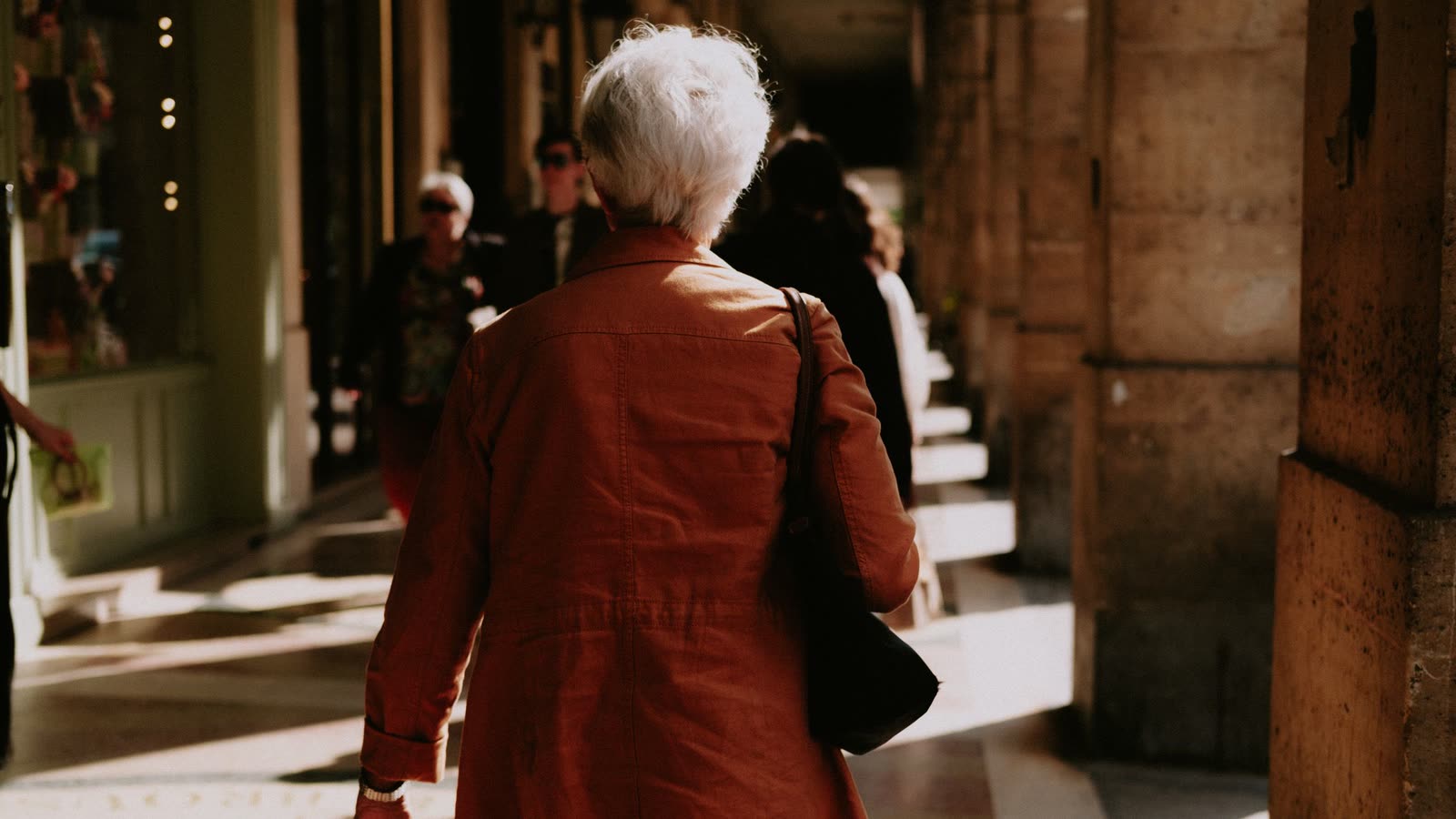 Femme âgée aux cheveux gris courts, vue de dos, marchant d'un pas assuré dans une galerie ensoleillée, chiropratique et prévention des chutes à Toulouse Nord